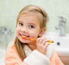 Child brushing teeth with an orange toothbrush in a bathroom, smiling toward the camera.