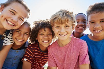Five smiling children leaning close together outdoors under a bright sky.