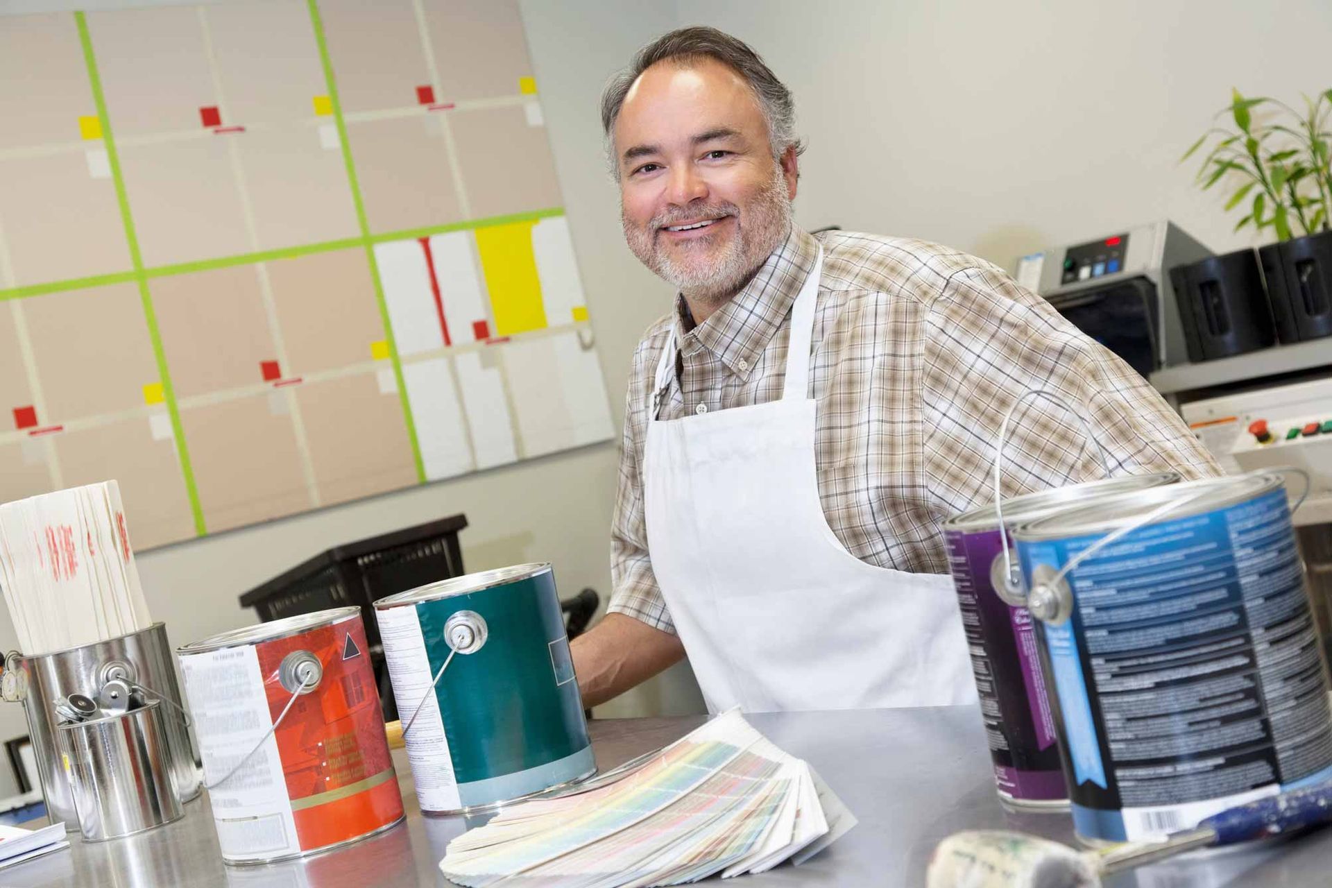 Smiling man in a white apron painting at a worktable with colorful cans in a studio