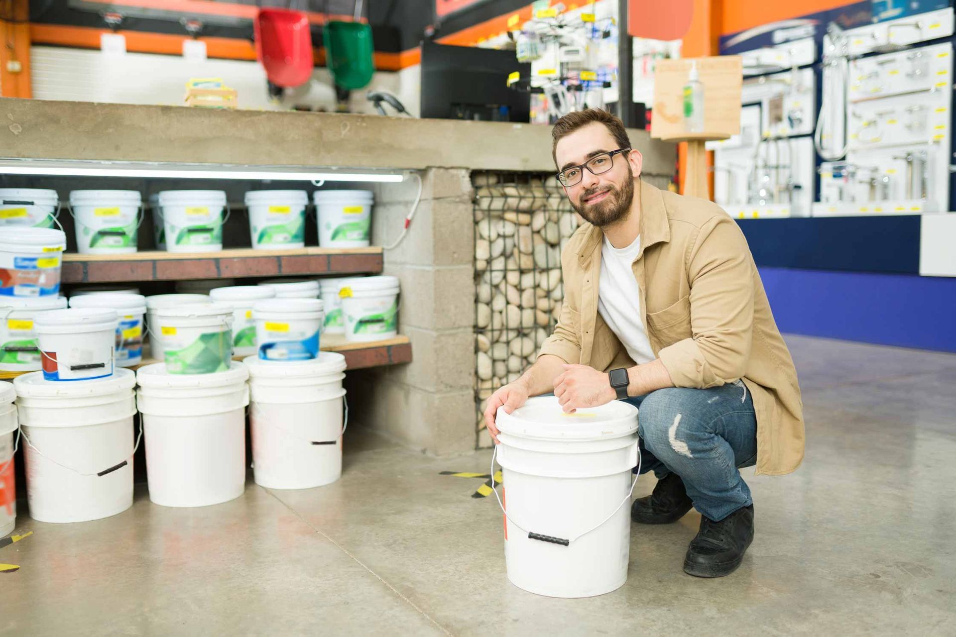Man crouching in a storage aisle beside stacked white buckets and paint buckets on shelves