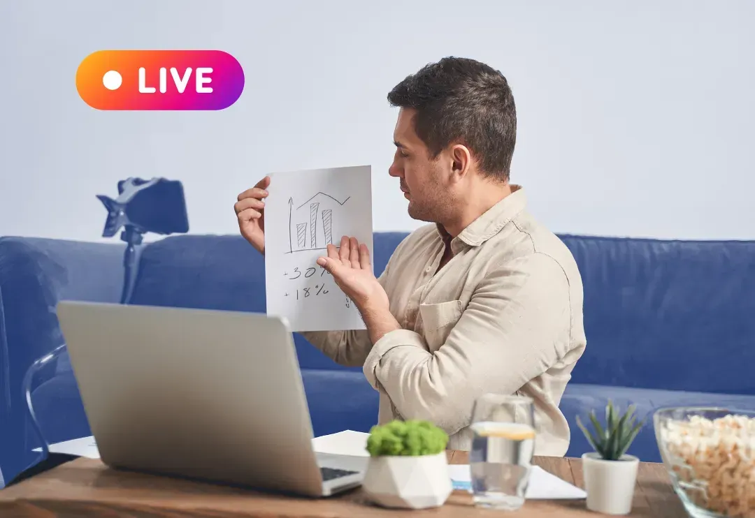 A man is sitting in front of a laptop computer holding a piece of paper.