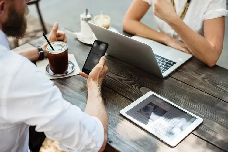 A man is holding a cell phone while sitting at a table with a laptop and a tablet.