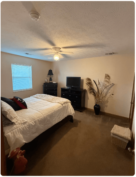 Cozy bedroom with unmade bed, dresser, TV, ceiling fan, window blinds, and vase of dried branches.