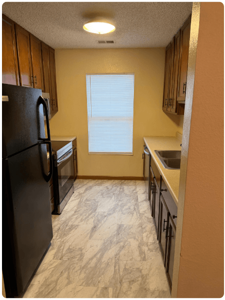 Narrow kitchen with black appliances, wood cabinets, beige walls, and a window at the end.