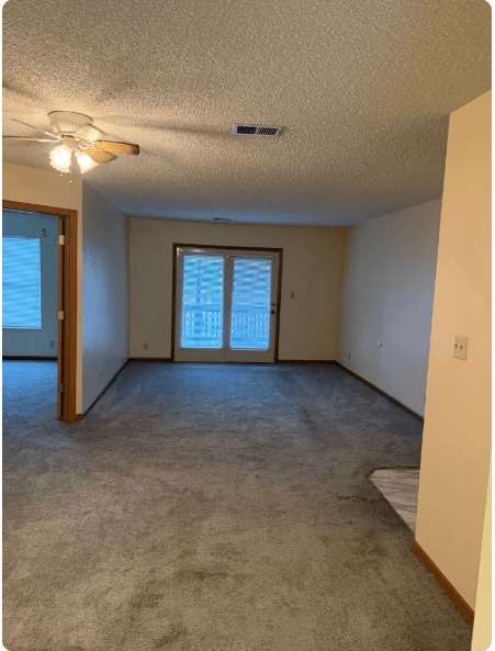 Empty carpeted living room with ceiling fan, sliding glass doors, and beige walls