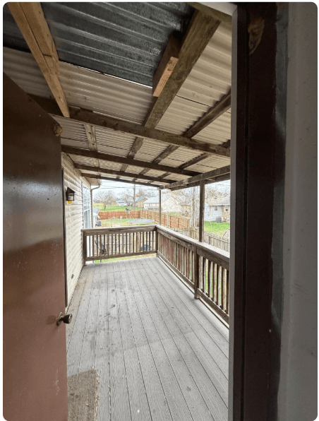 Covered wooden porch with railing and corrugated metal roof, viewed through an open doorway.