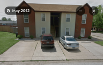 Apartment building entrance with two parked cars in front, seen from the street.