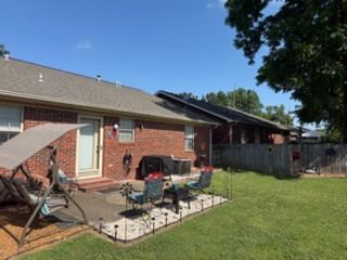 Backyard patio with chairs, grill, and fenced lawn beside a brick house under a clear blue sky