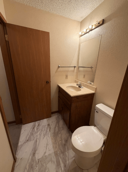 Small bathroom with toilet, vanity sink, mirror, wood door, and marble-pattern floor.