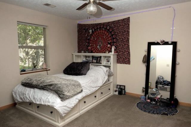 Bedroom with a white bed, patterned wall hanging, window, ceiling fan, and standing mirror.