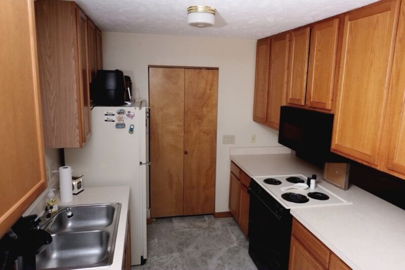 Narrow kitchen with wooden cabinets, white fridge, sink, stove, and microwave along both walls.