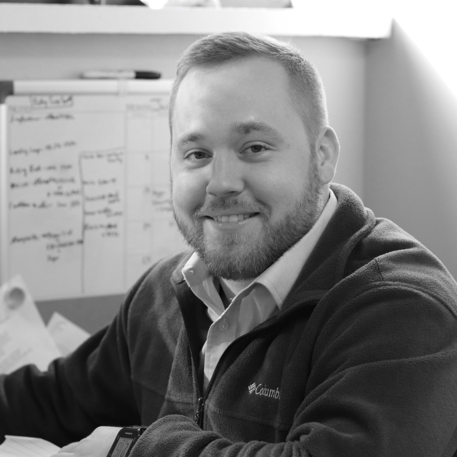 A man with a beard is smiling in a black and white photo while sitting at a desk.