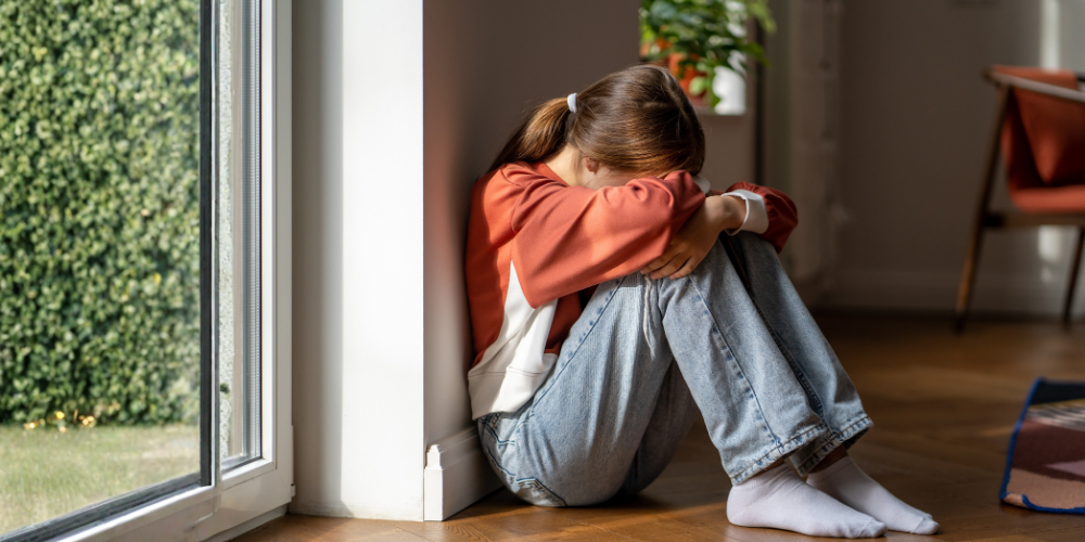 Child sitting on the floor by a window, hugging knees and hiding face, in a dim room