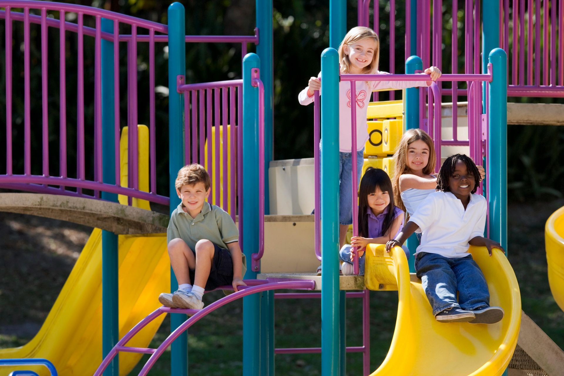 Children on a colorful playground, sitting and sliding on yellow slides with purple railings.