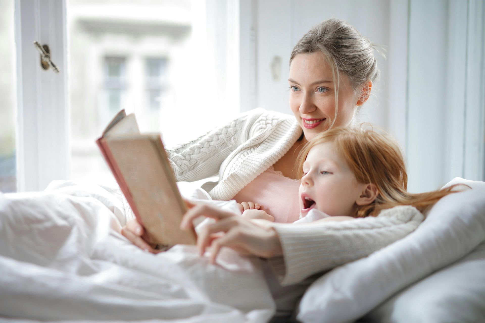 A person reads a book to a child while they both lie comfortably in a bed near a window.