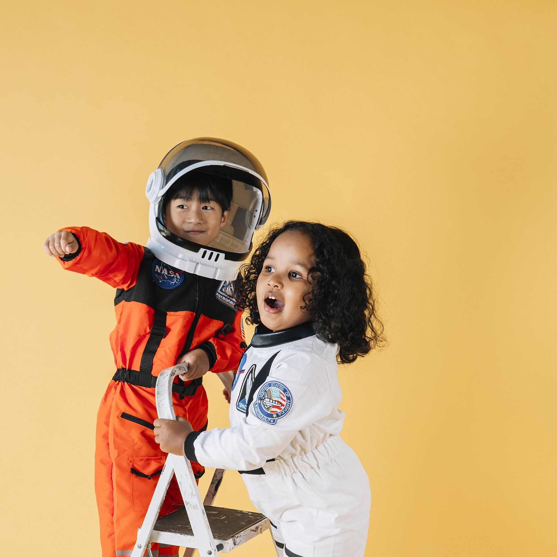 Two children wearing astronaut suits stand by a white ladder against a plain yellow background.