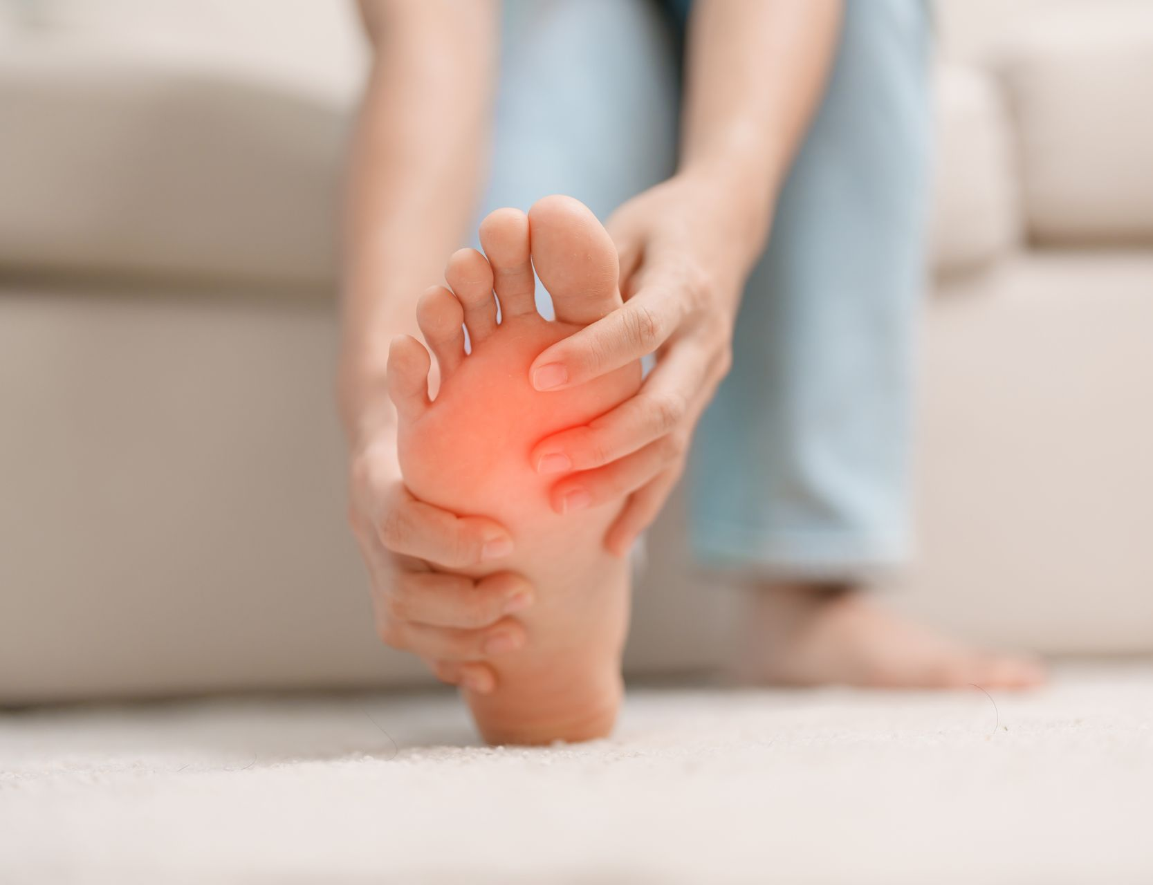 Person holding foot, showing pain area highlighted in red. Sitting on a couch, indoors.