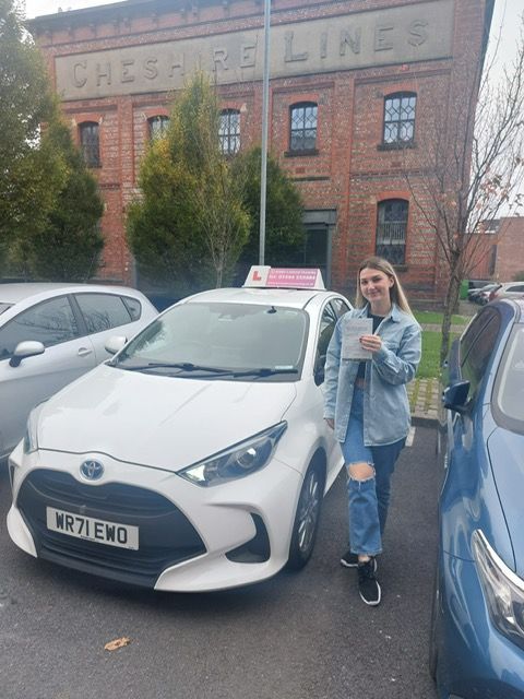 A woman is standing next to a white car in front of a brick building.