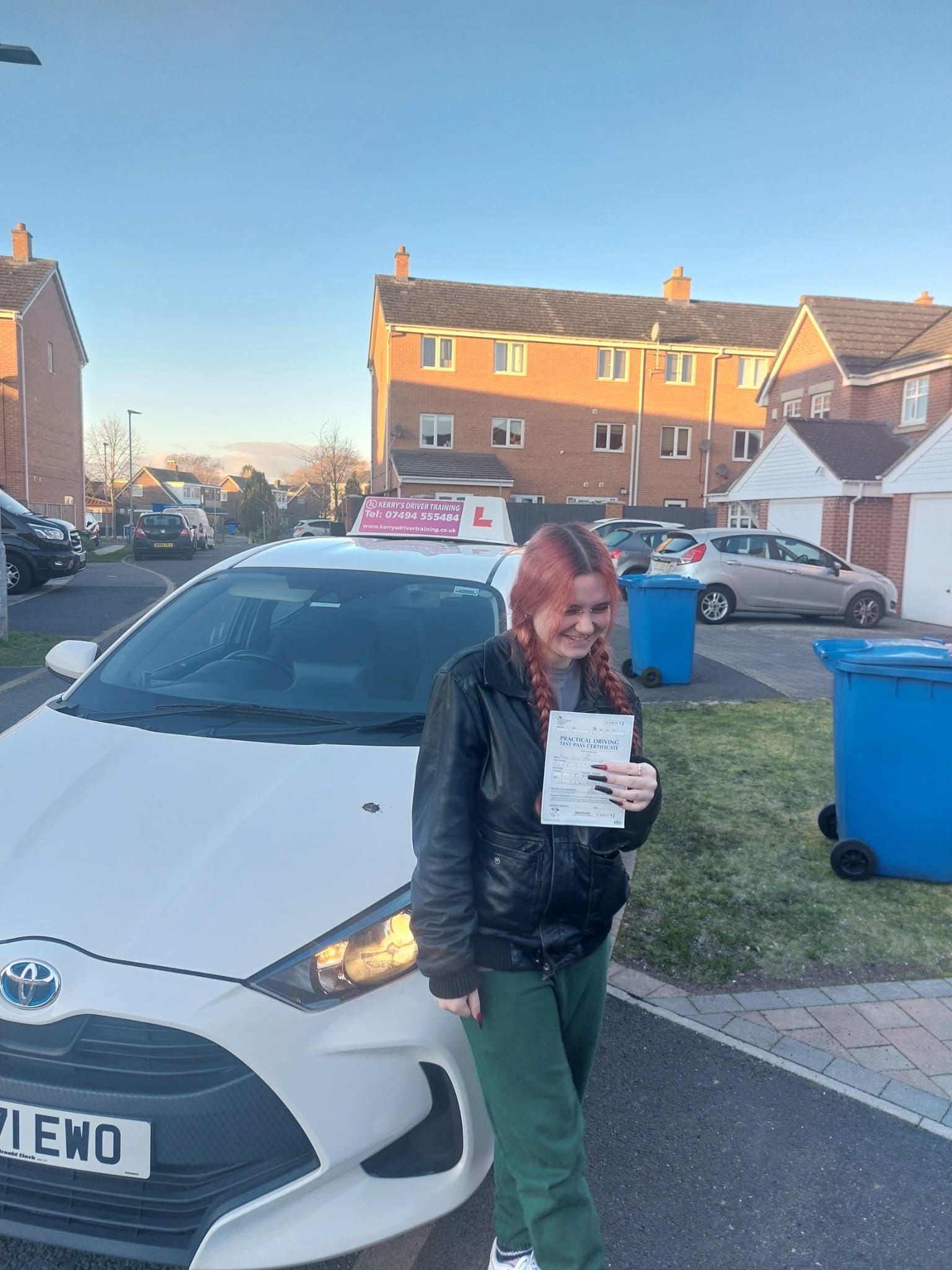 Woman holding driving test pass certificate, smiling in front of white car with