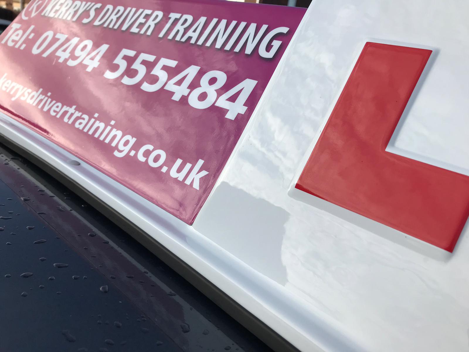 Close-up of a white learner driver car sign with a red L and training text on a pink banner.