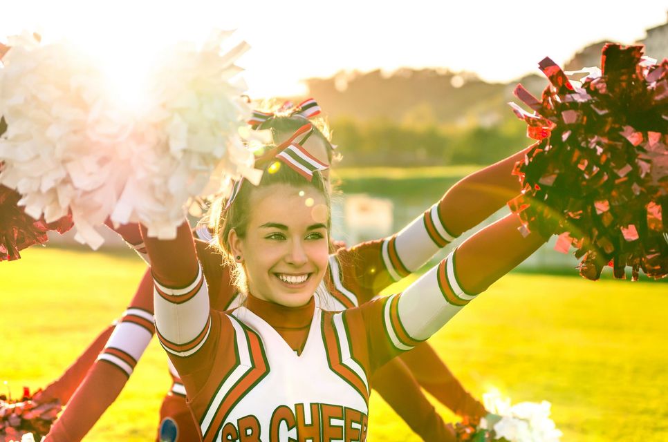 Cheerleaders in orange and white uniforms with pom-poms cheering in sunlight.