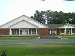 Single-story brick and white building with columns, set behind a parking lot and green lawn