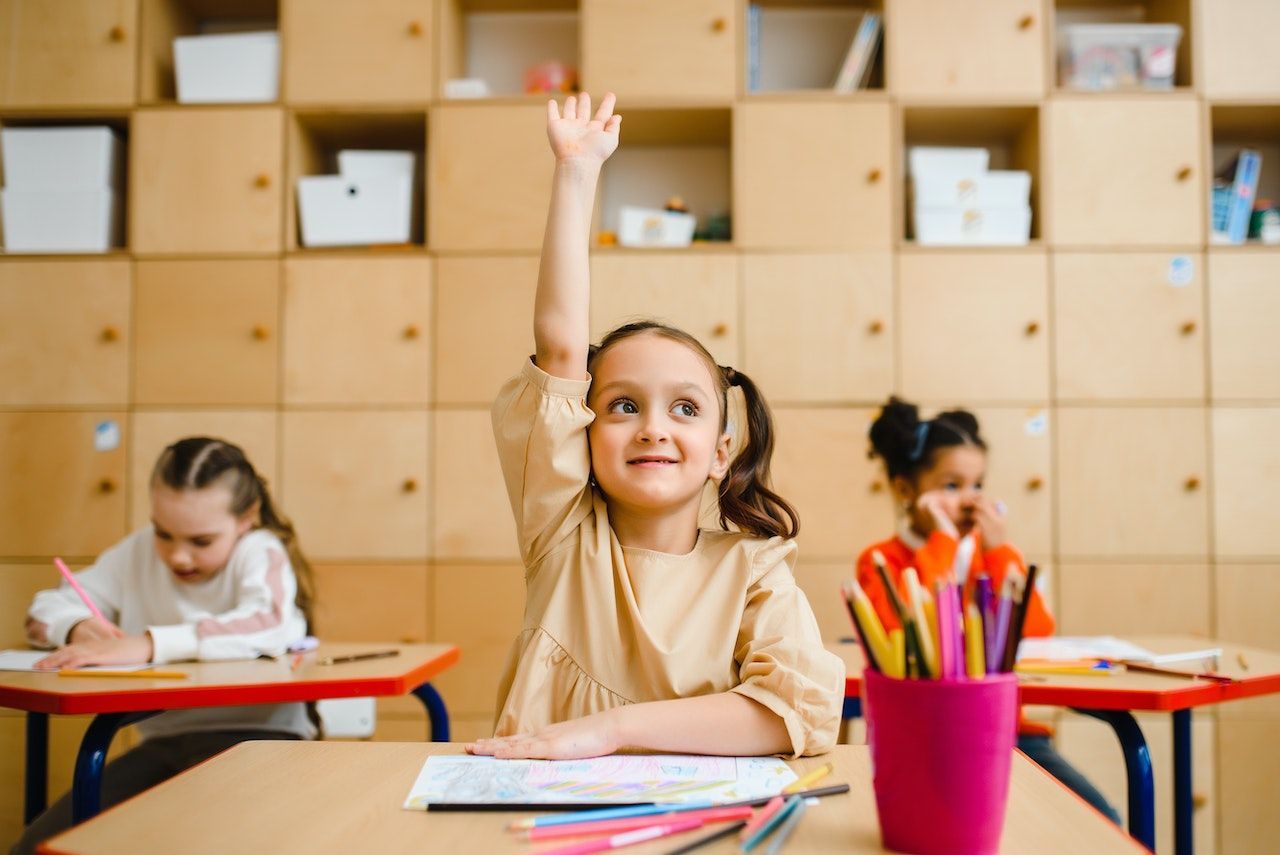 A little girl is raising her hand in a classroom to answer a question.