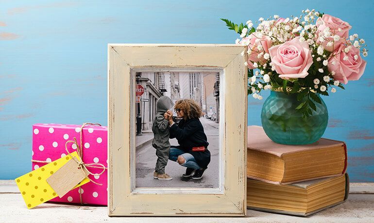 Framed photo of a child with an adult beside pink flowers, wrapped gifts, and stacked books on a table