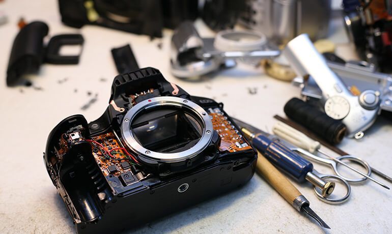 Broken black camera with lens cap off, surrounded by scattered repair tools on a workbench.
