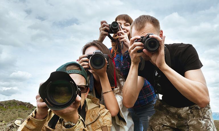 Group of photographers crouching outdoors, aiming cameras toward the viewer under a cloudy sky