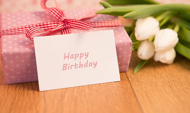 Happy Birthday card beside a pink gift box with red ribbon and white tulips on a wooden table