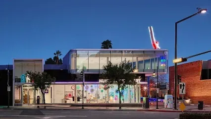 Glowing neon art installations visible through MONA's glass walls at night, vibrant pinks and blues illuminating bustling visitors inside