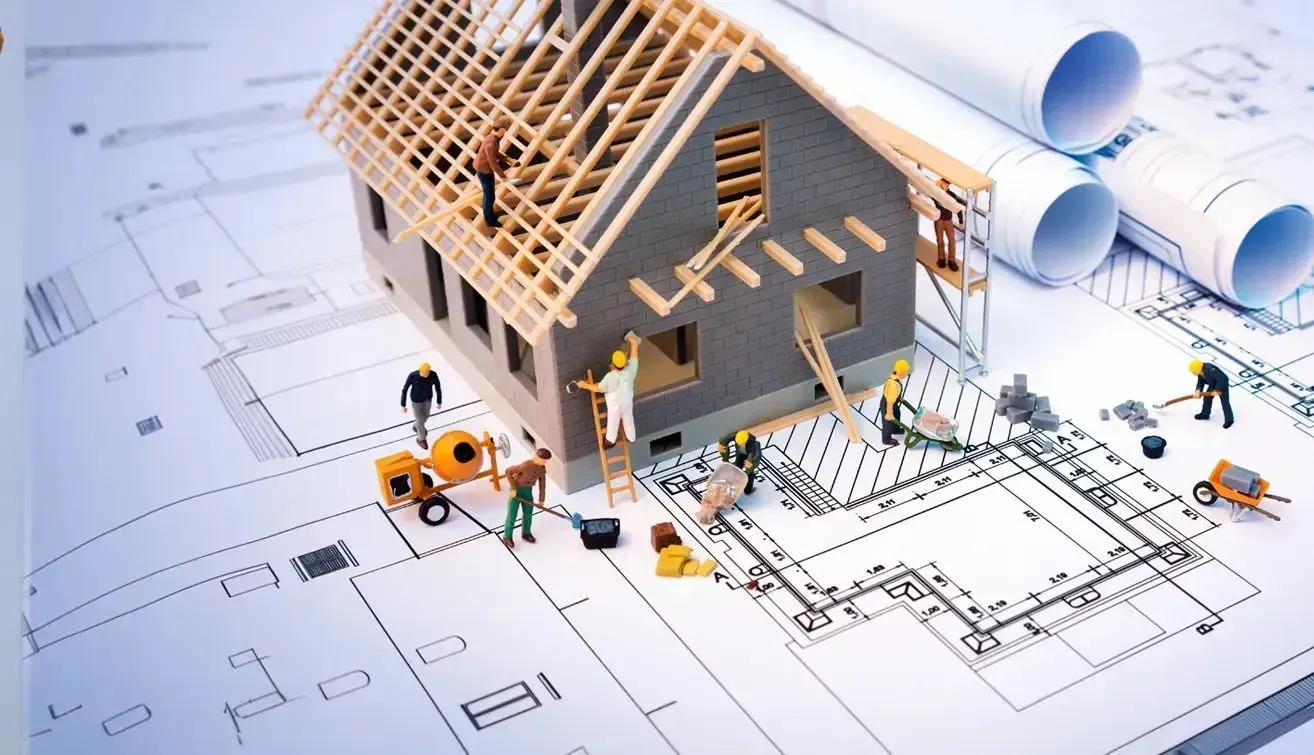 Miniature construction site with tiny workers fixing a dollhouse-sized building, surrounded by a rolled-out paper blueprint