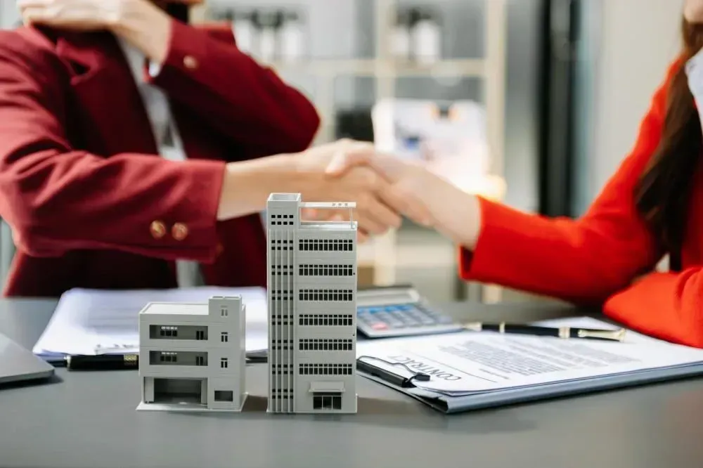 Real estate agent and client shaking hands across a table with signed documents, sealing a property deal