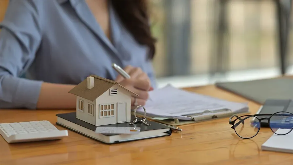 Businesswoman in blue polo signing contract at table, with miniature house model, magnifying glass, and notebook placed strategically—symbolizing meticulous property review