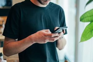 A person in a black t-shirt uses a smartphone while standing near a window with a green plant in the foreground.