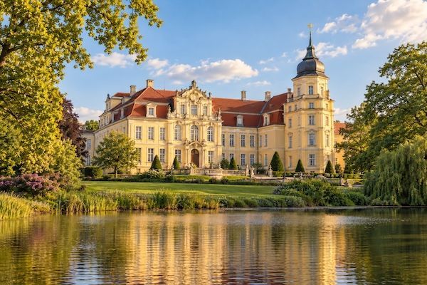 Illustratives Schlossmotiv für Oldenburg mit heller Fassade, Park und Spiegelung im Wasser
