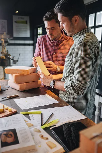 Dois homens estão de pé em uma mesa olhando para um pedaço de queijo.