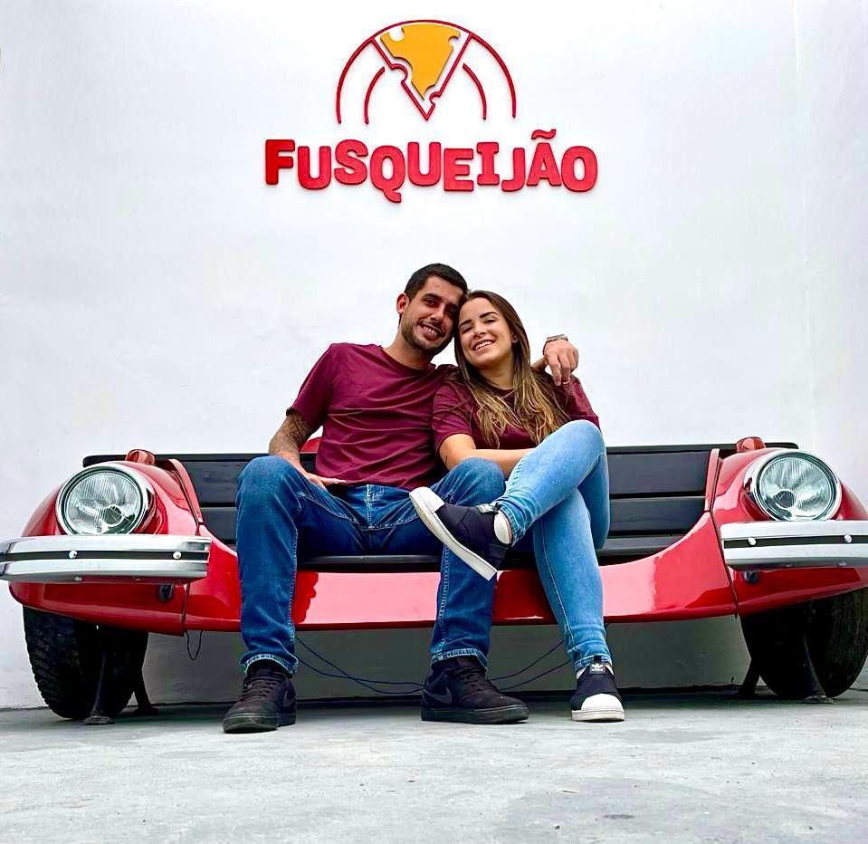 Um homem e uma mulher estão sentados em um banco em frente a uma placa que diz fusqueijao