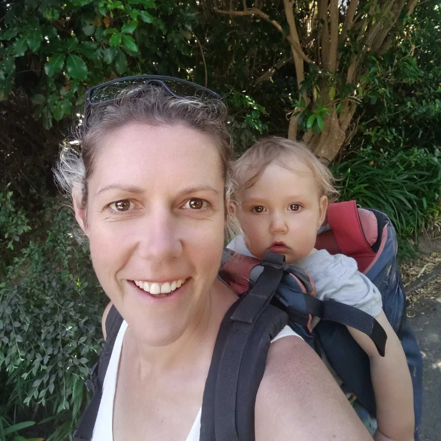 Smiling woman carrying a baby in a backpack carrier outdoors среди greenery