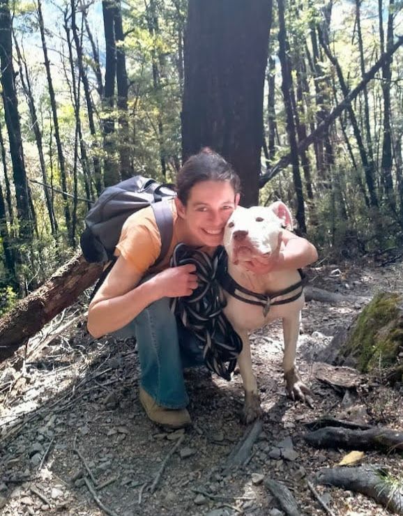 Hiker kneels with a white dog on a forest trail, both facing the camera in dappled sunlight