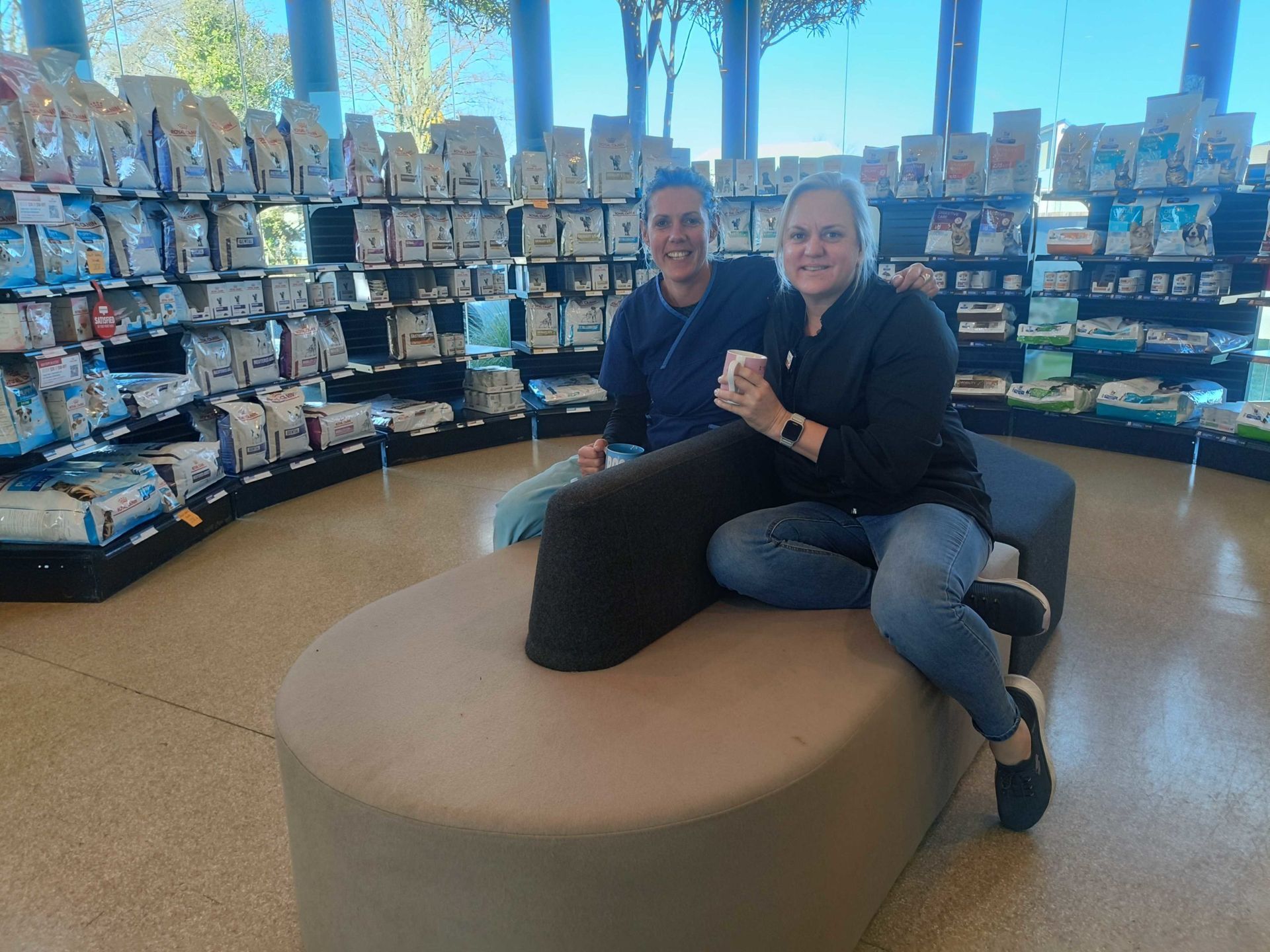 Two people sit smiling on a round tan bench in a store with shelves of books and cards behind them.