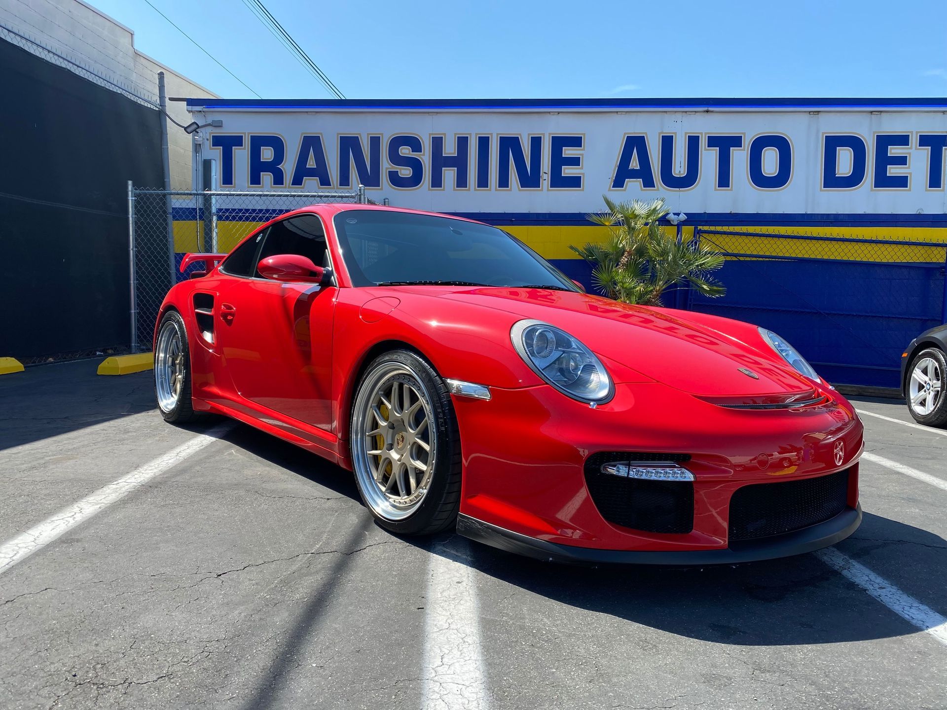A red porsche 911 turbo gt2 is parked in a parking lot in front of a transshine auto detailing shop.