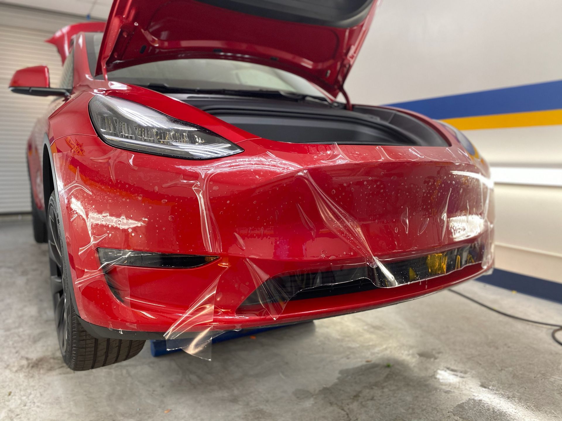 A red tesla model 3 with its hood up is covered in plastic.
