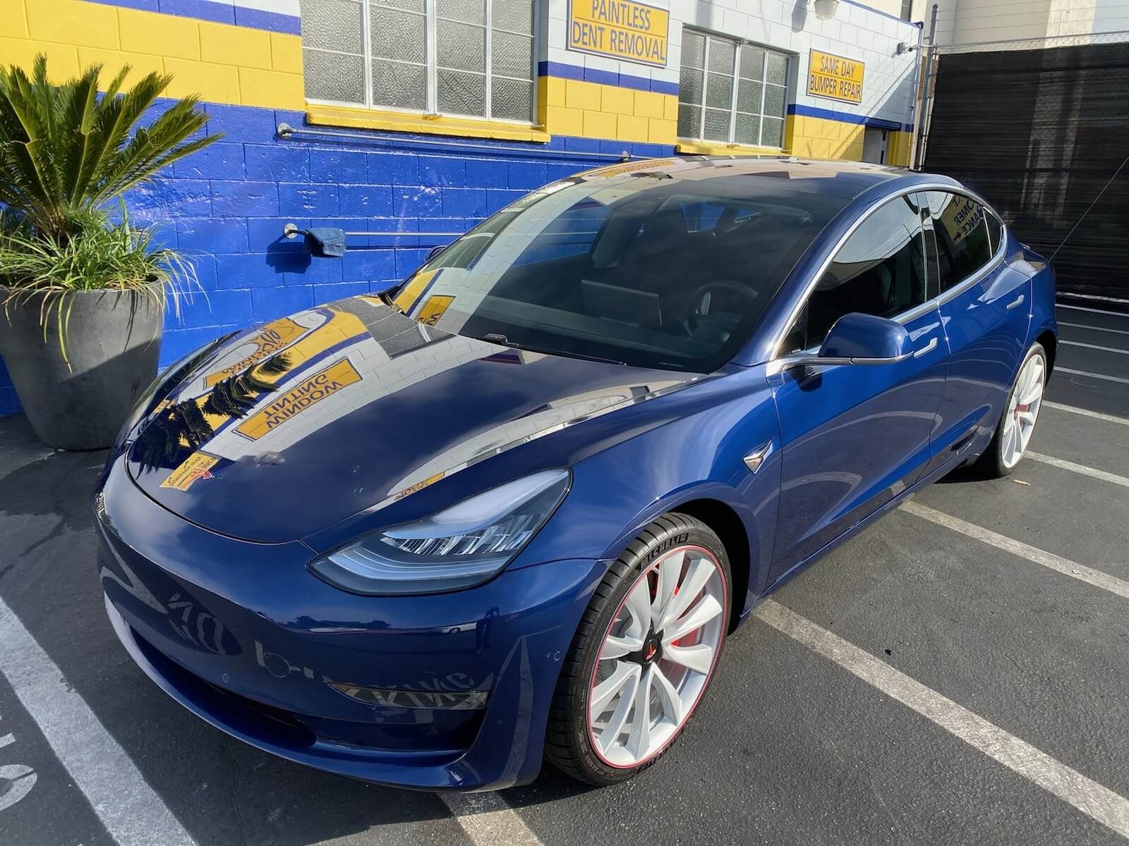 A blue tesla model 3 is parked in a parking lot in front of a building.