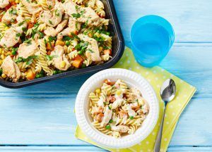A bowl of chicken pasta next to a baking dish of the same, a blue cup, and a spoon on a light blue wooden surface.