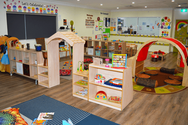 A brightly lit preschool classroom with wooden tables, chairs, and open shelving units filled with various toys.