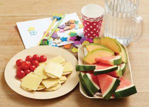 A plate of cheese and crackers, a bowl of watermelon and melon slices, a polka-dot cup, and craft supplies on a table.