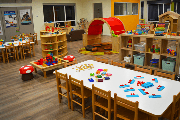 A brightly lit preschool classroom with tables, chairs, toy shelves, and a small play tunnel on a wooden floor.
