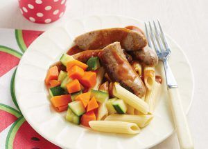 A plate containing cooked penne pasta, sausages in a savory sauce, and a side of diced carrots and zucchini.